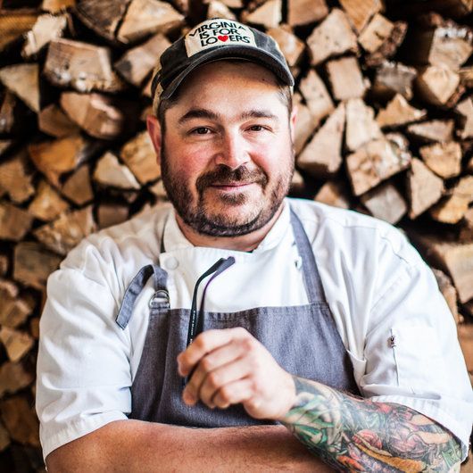 a man wearing an apron and hat standing in front of stacks of wood with his arms crossed