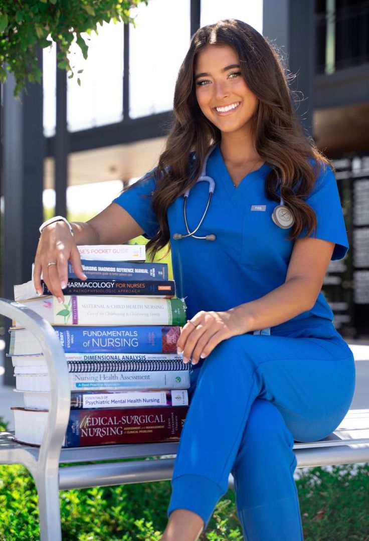 a woman in blue scrubs sitting on a bench with several books stacked on her lap