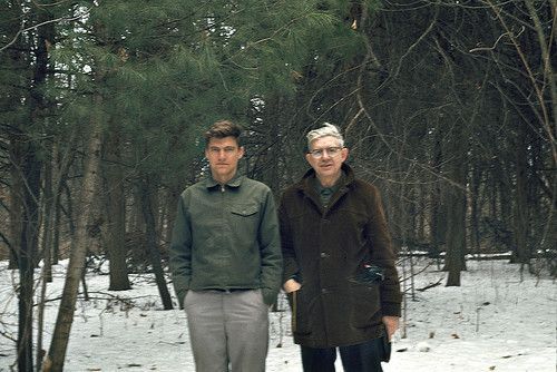 two people standing next to each other in the snow with trees and bushes behind them