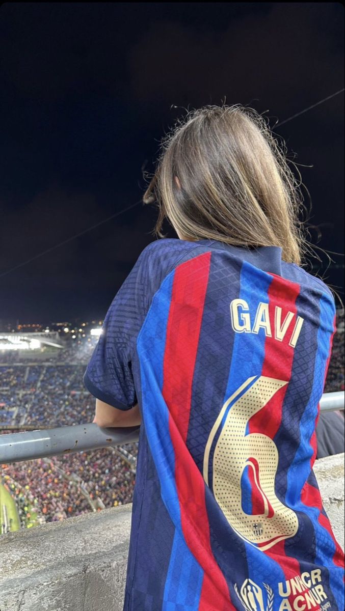 a woman in a blue and red shirt is looking at the soccer field while holding a cell phone