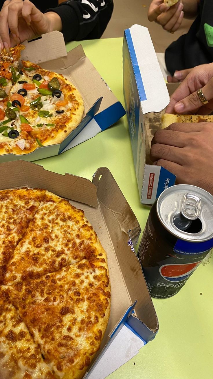 three people eating pizza and drinking soda at a table with two boxes of pizza in front of them