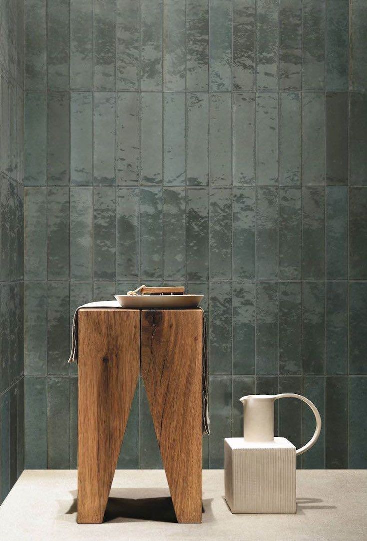 a wooden table sitting next to a white vase on top of a counter in front of a green tiled wall