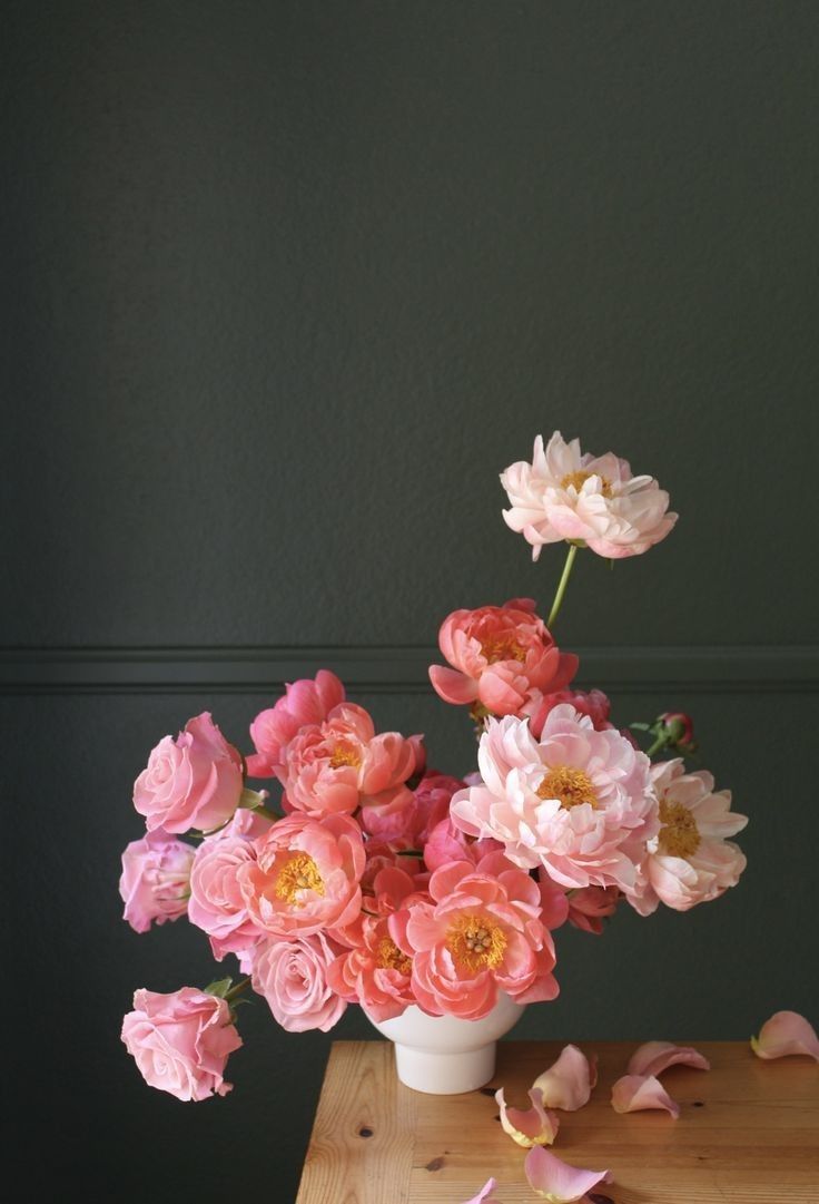 a white vase filled with pink flowers on top of a wooden table next to a black wall