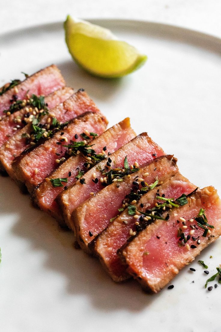 slices of meat on a white plate next to a lime and some seasoning sprigs