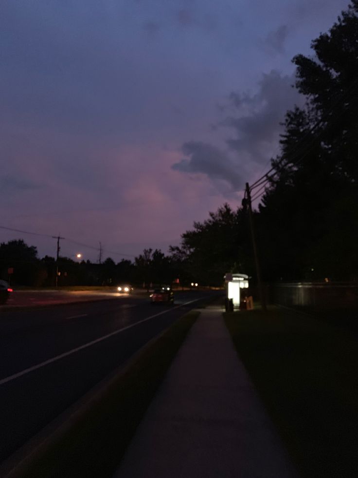 an empty sidewalk next to a street at night with cars driving on the road and dark clouds in the sky