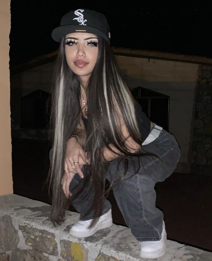 a woman with long hair sitting on top of a stone wall wearing a baseball cap