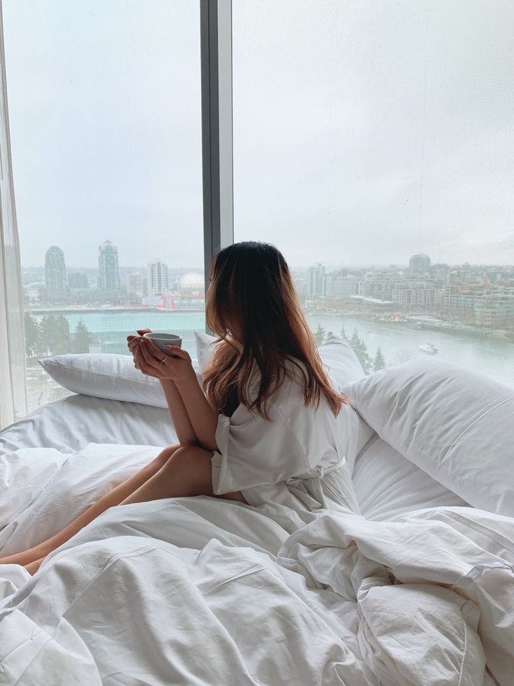 a woman sitting on a bed looking at her cell phone in front of a window