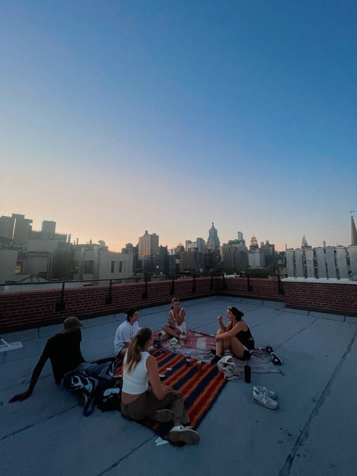 four people sitting on top of a roof eating pizza and drinking beer at sunset or dawn