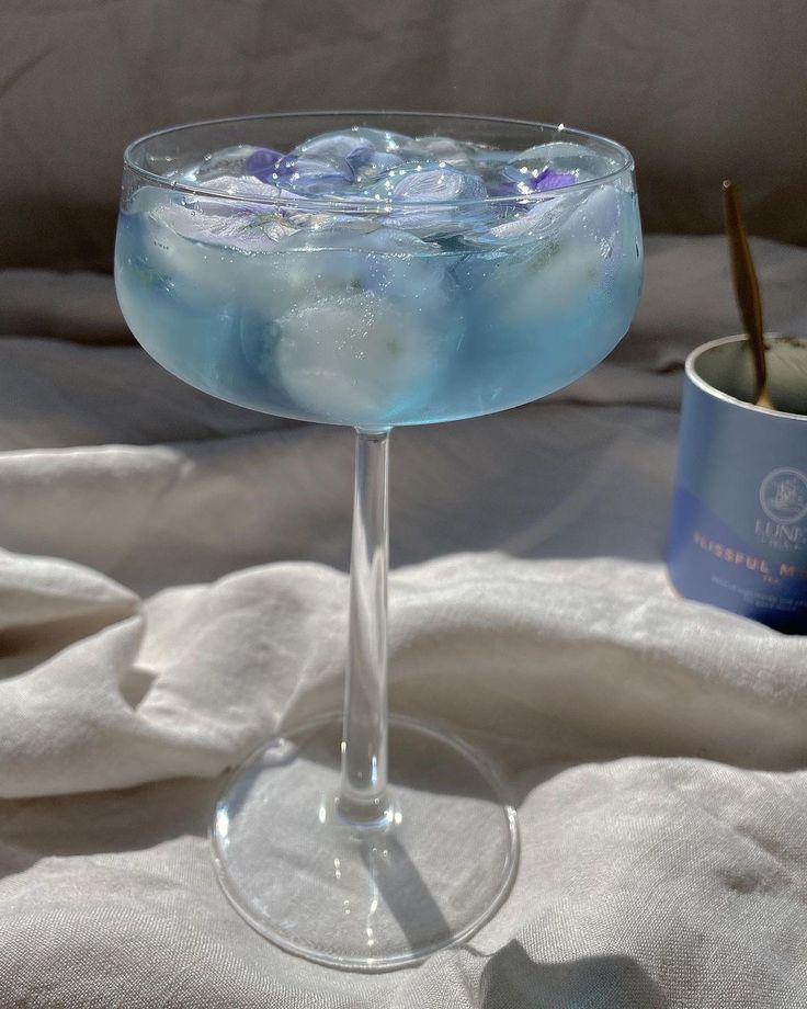 a glass filled with blue liquid sitting on top of a white blanket next to a cup