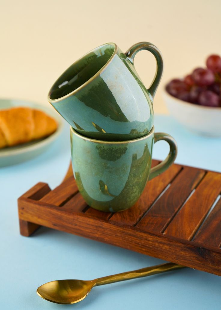 two green coffee cups sitting on top of a wooden tray next to grapes and croissants