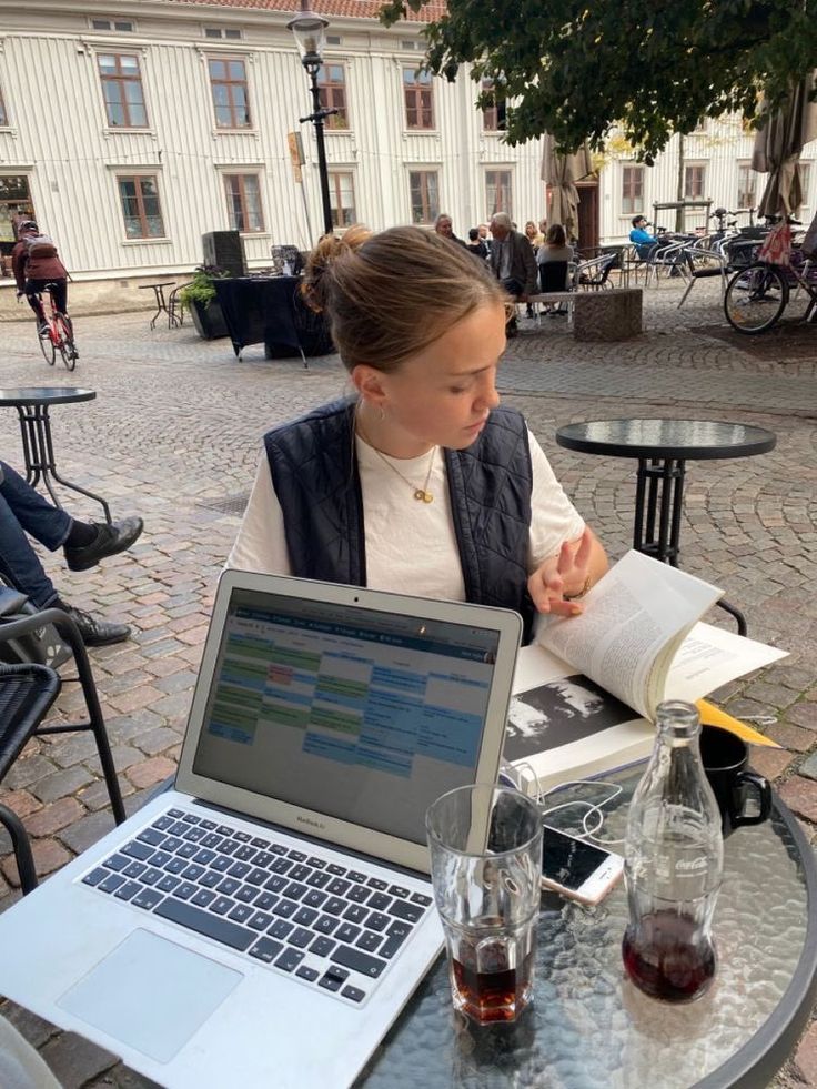 a woman sitting at an outdoor table with a laptop and book in front of her