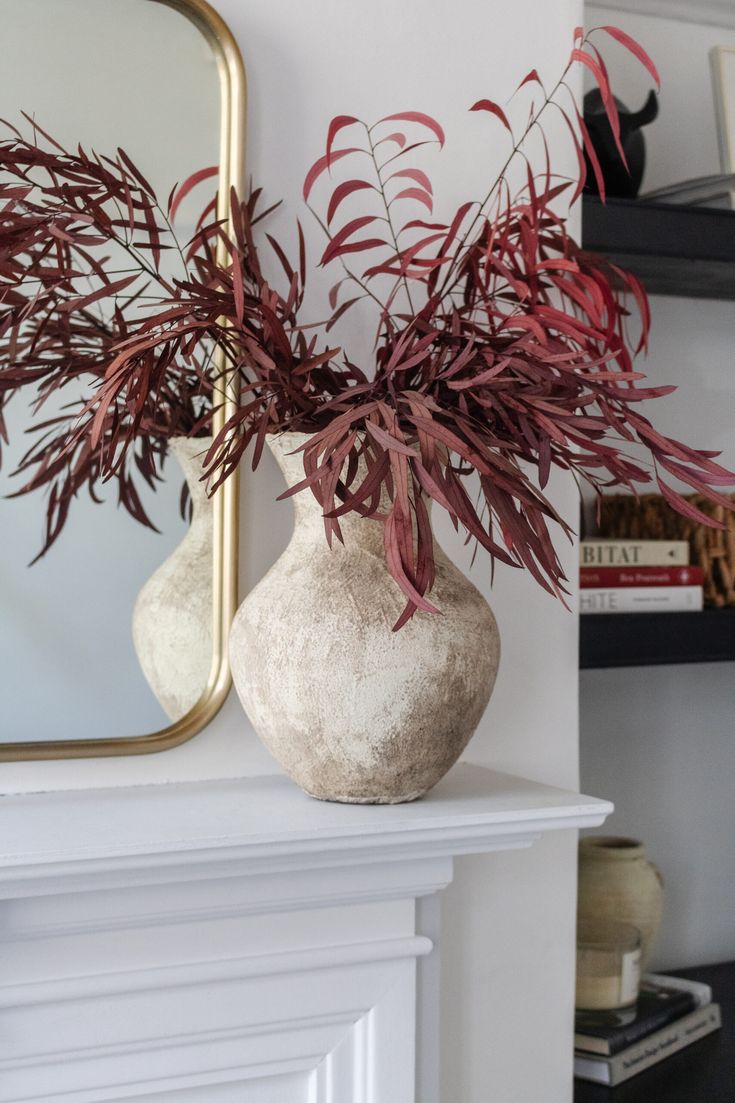 a vase with some red plants in it next to a mirror on a shelf above a fireplace