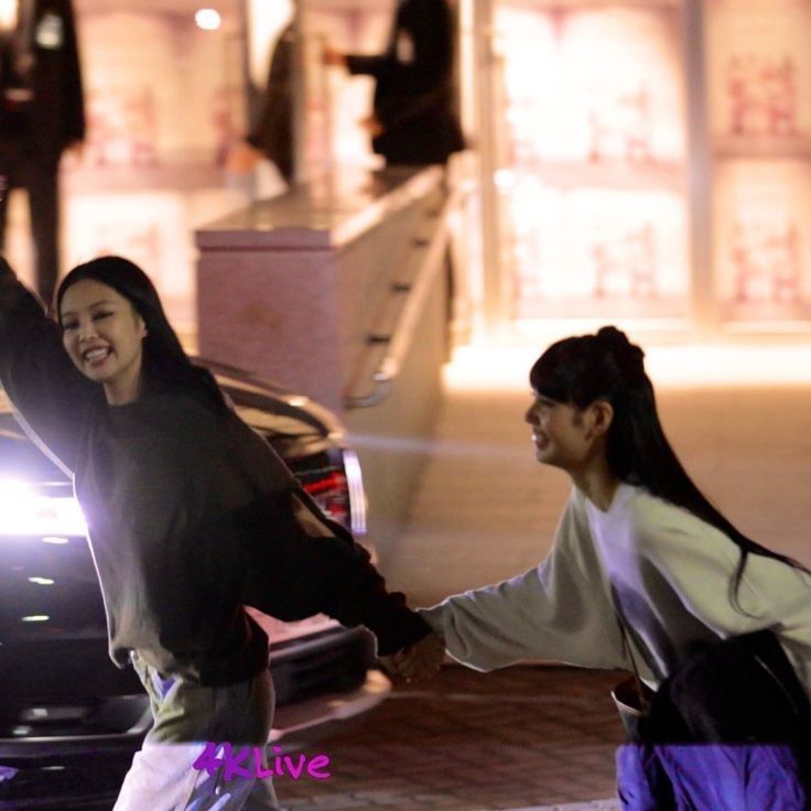 two young women holding hands and walking down the street at night with their arms in the air