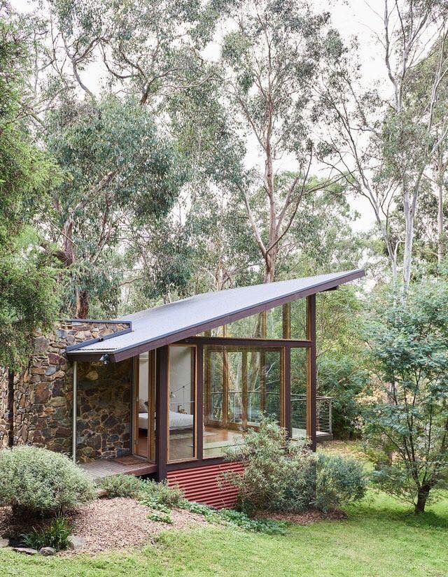 a small house in the middle of some trees and grass with a stone wall behind it