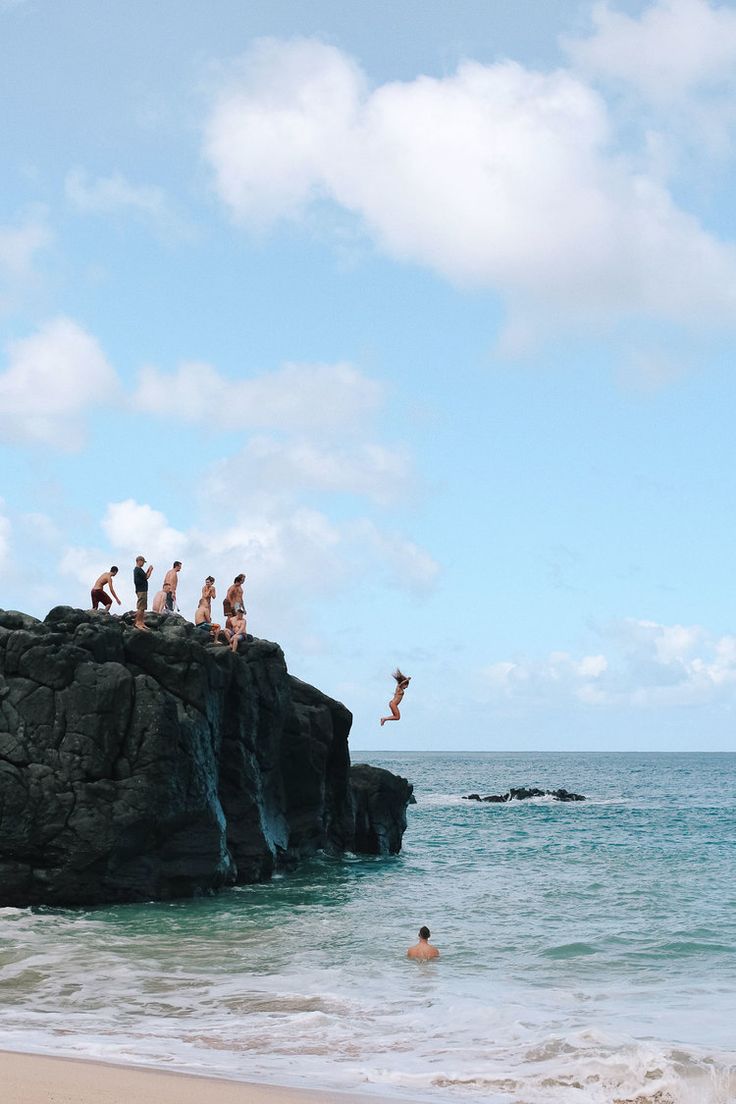 people are jumping off the rocks into the water at the beach while another person swims in the ocean