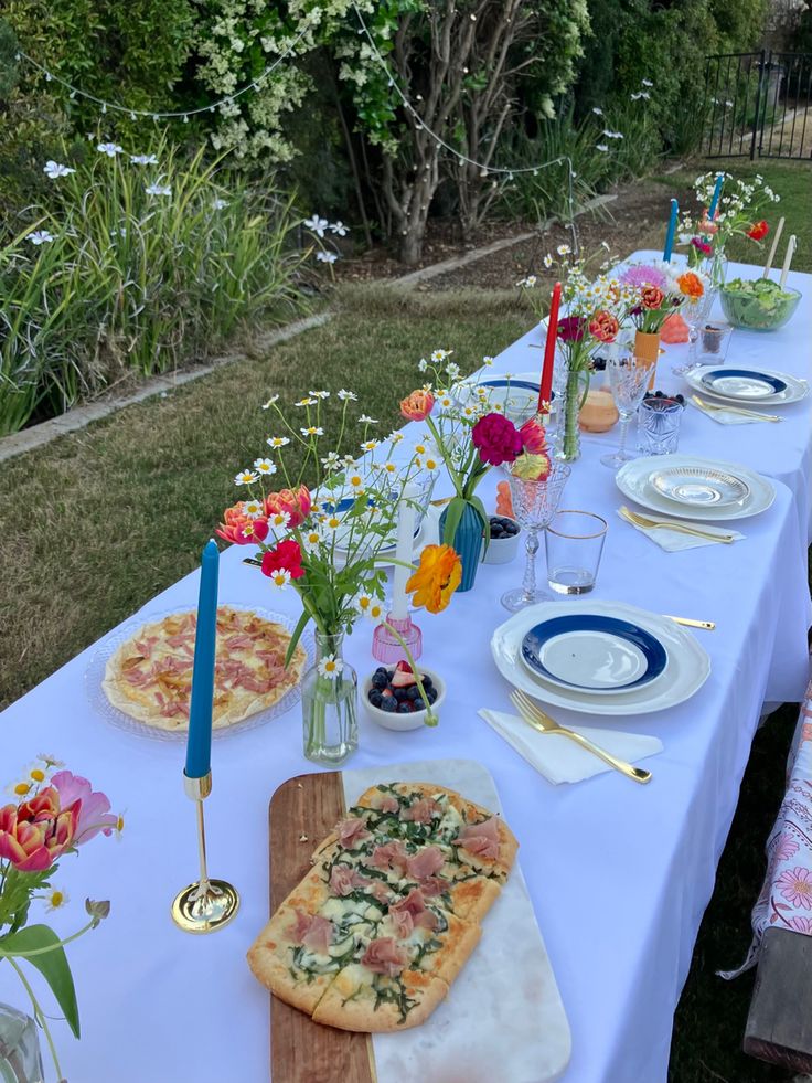 a long table is set with pizza, flowers and candles for an outdoor dinner party