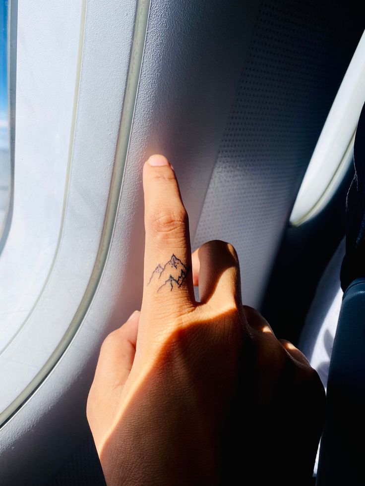 a person's hand with a small tattoo on it pointing to the sky through an airplane window