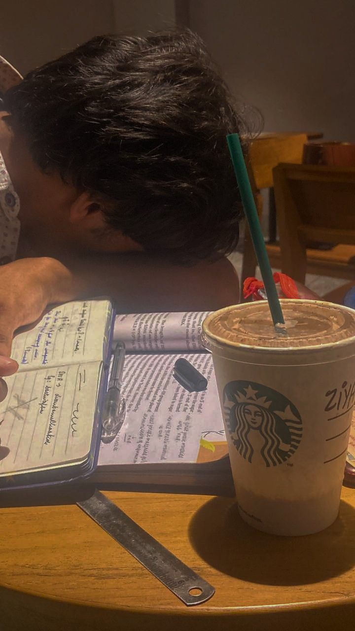 a person writing on a notebook next to a cup of coffee with a straw in it