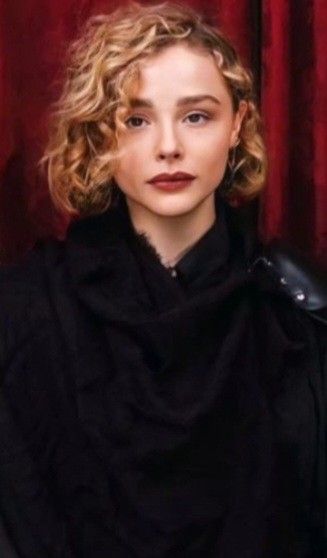 a woman with curly hair is wearing a black coat and standing in front of a red curtain