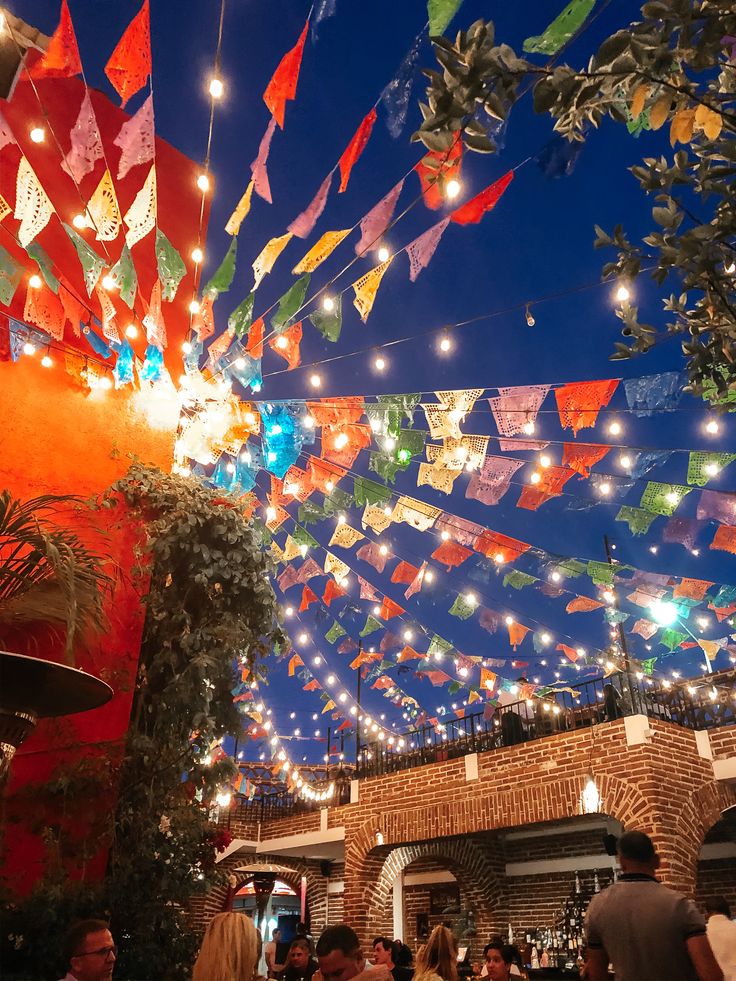 many colorful flags are hanging from the ceiling above people at an outdoor bar and restaurant