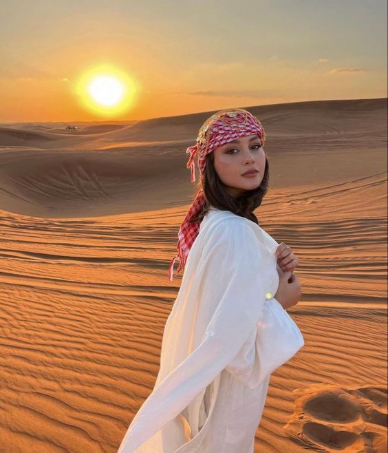 a woman standing in the desert with her arms around her waist and head scarf draped over her shoulders