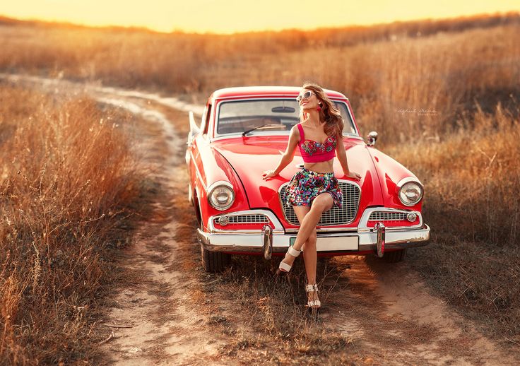 a woman sitting on top of a red car in the middle of a dirt road