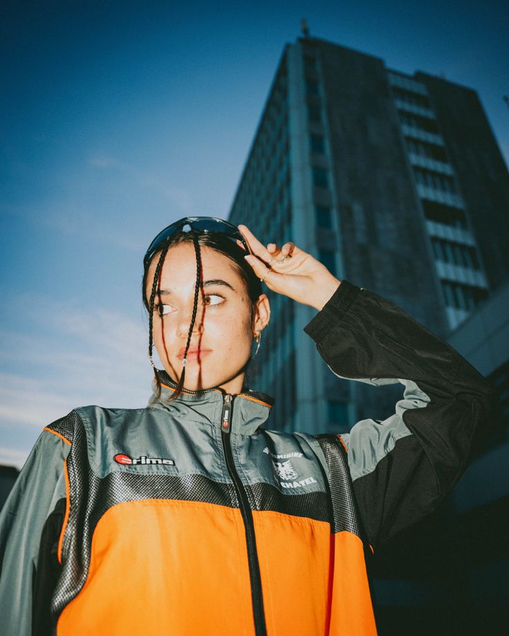 A girl in an orange racer jacket poses in front of a tall building. Racer Glasses, Nike Editorial, Building Photoshoot, Clean Girl Hairstyle, Sporty Photoshoot, Streetwear Photoshoot Ideas, Street Style Sporty, Hairstyles Brunette, Street Fashion Photoshoot