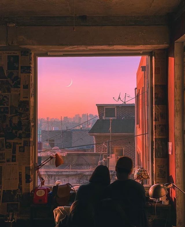 two people looking out an open window at the city below and pink sky in the distance