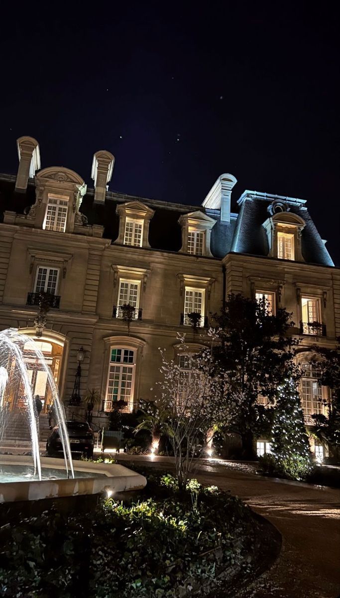 a fountain in front of a large building at night with lights on the windows and trees around it