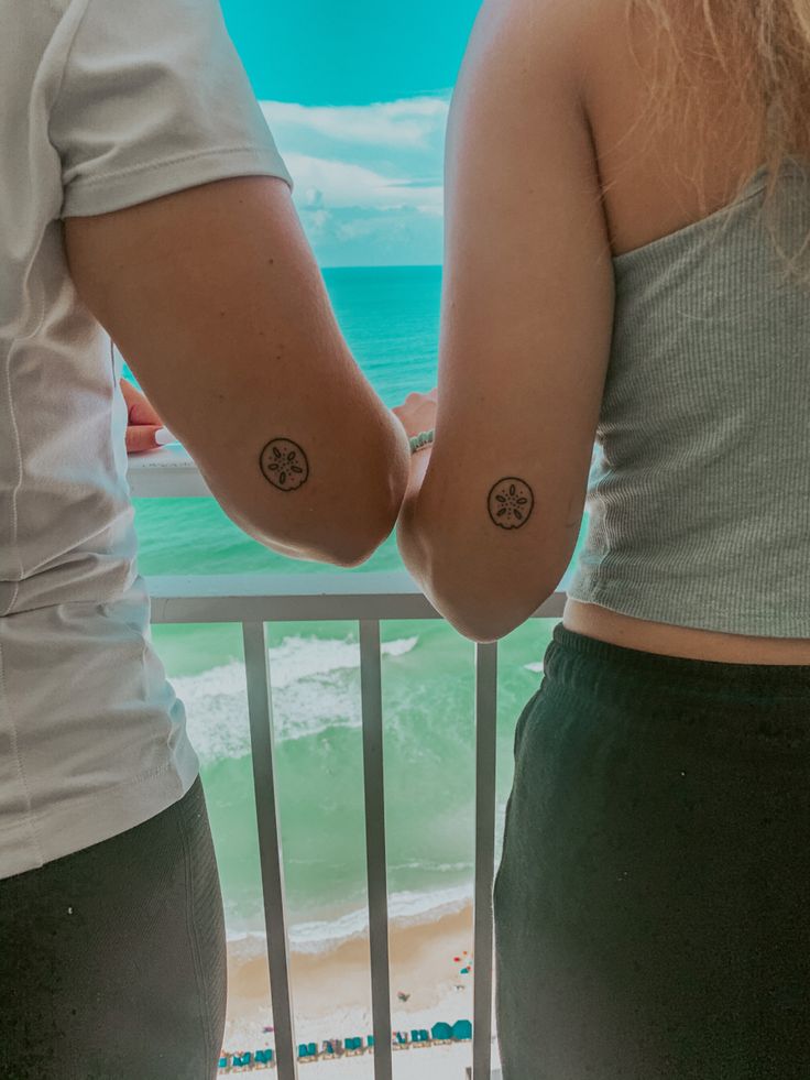 two people standing next to each other with tattoos on their arms and shoulders, facing the ocean