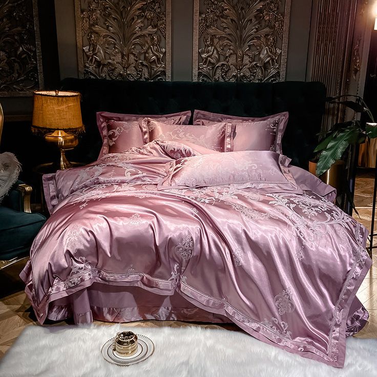 a bed with pink comforter and pillows in a room next to a lamp on a table