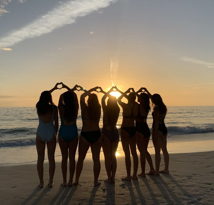 six women in bathing suits standing on the beach at sunset with their hands up to heart shape