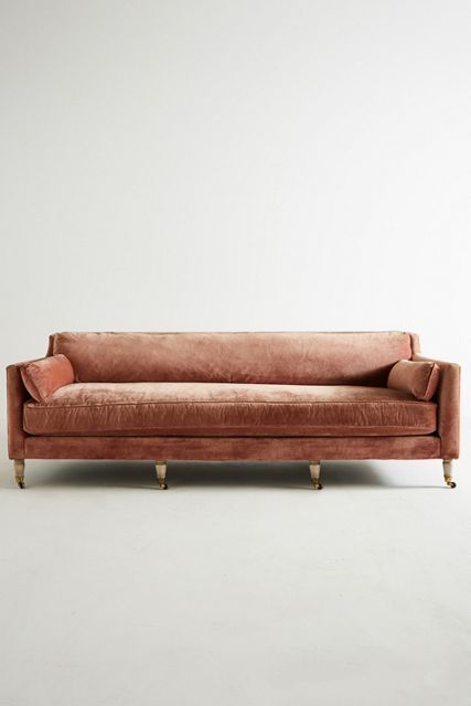 an orange velvet couch with gold legs and pillows on the back, in front of a white wall