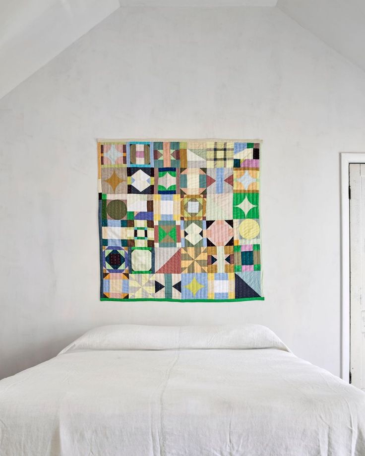 a bed with a quilt hanging on the wall above it in a room that has white walls