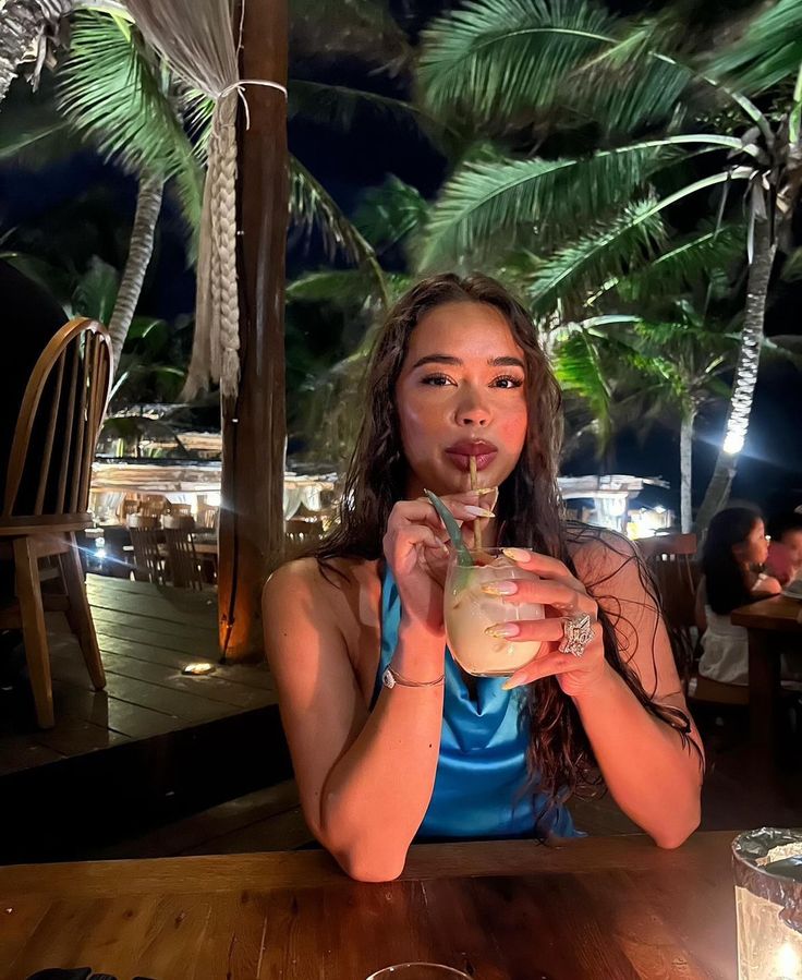 a woman sitting at a table with a drink in her hand and palm trees behind her