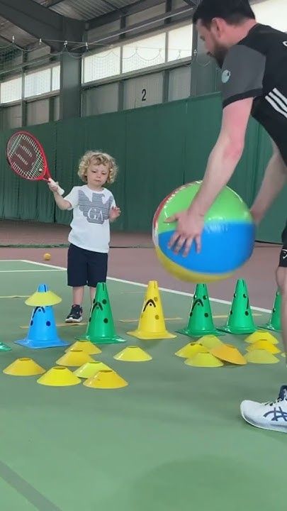 a man teaching a little boy how to play tennis with balls and rackets on an indoor court