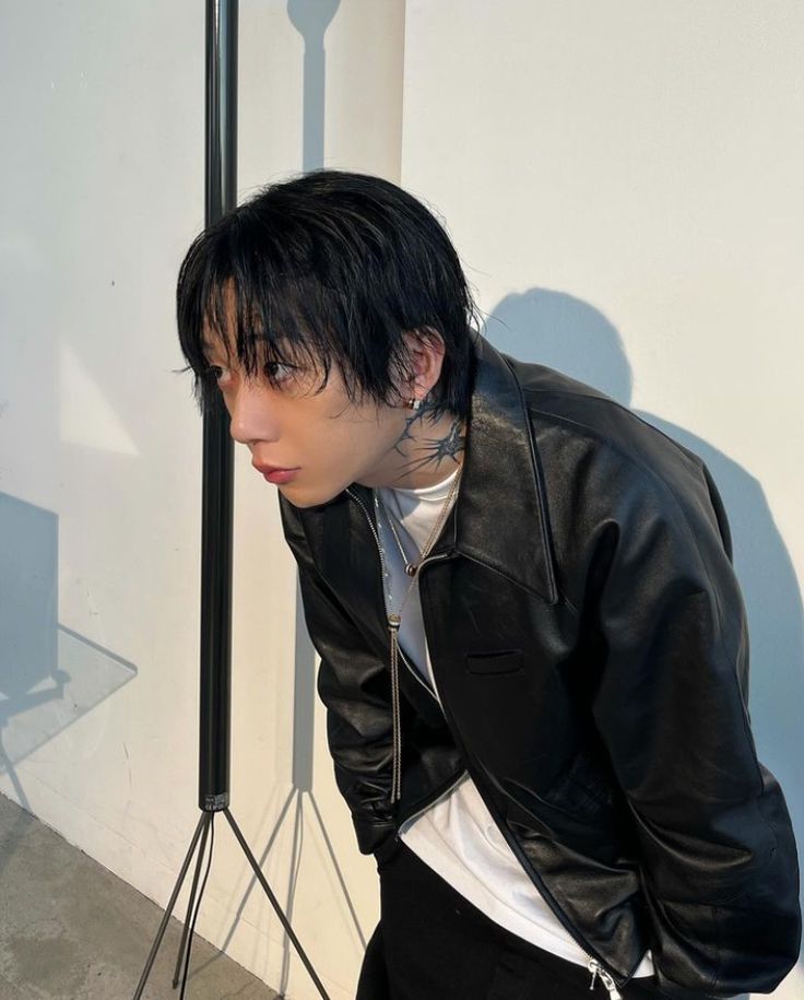 a man with black hair and piercings wearing a leather jacket standing in front of a white wall