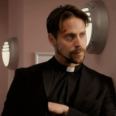 a man in a priest's outfit standing next to a wall mounted mirror and looking at the camera