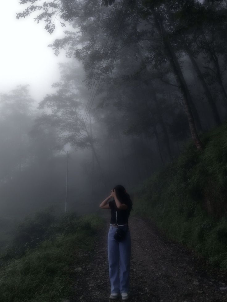 a woman is standing on a foggy path in the woods with her hands behind her head