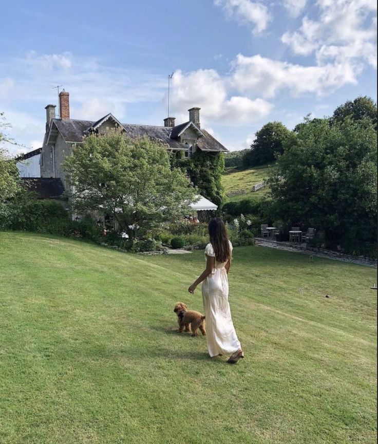 a woman in a white dress is walking her brown dog on the grass near a large house