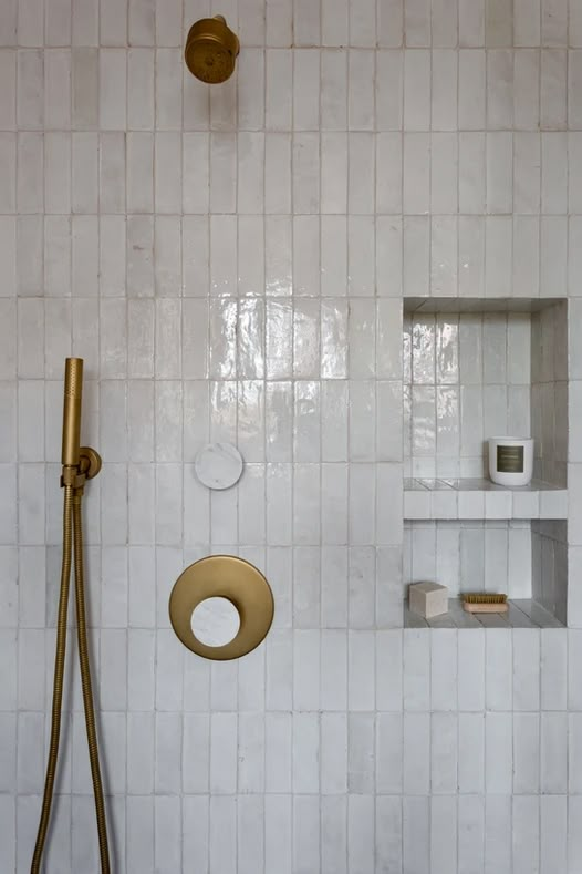 a white tiled shower with gold fixtures and shelves on the wall next to toilet paper