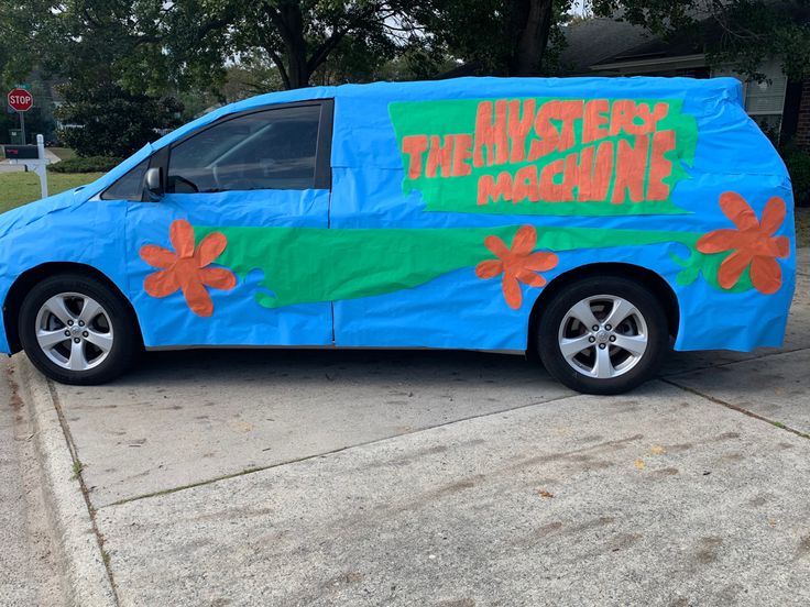 a blue van with flowers painted on it is parked in front of a house that says the mystery machine