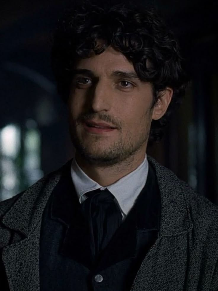 a man with curly hair wearing a gray coat and black tie in a dark room