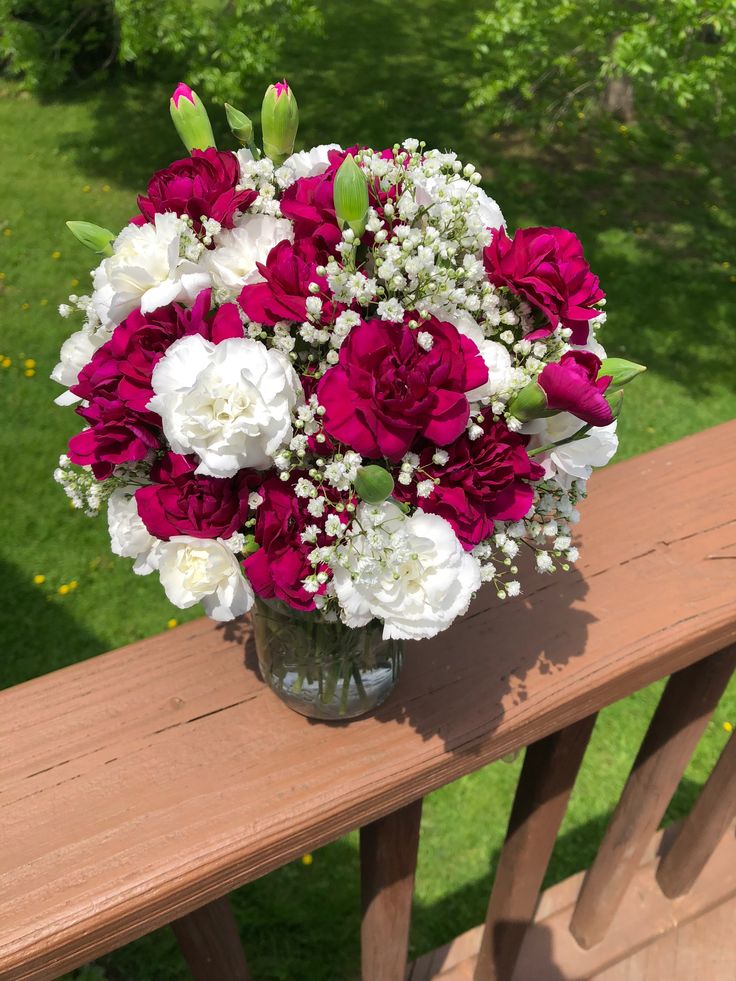 a vase filled with red and white flowers sitting on top of a wooden bench next to grass