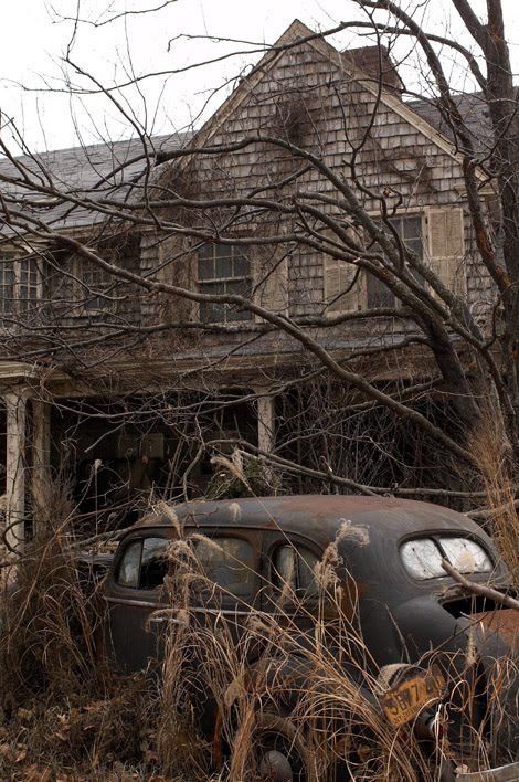 an old car is parked in front of a house that has been overgrown with weeds