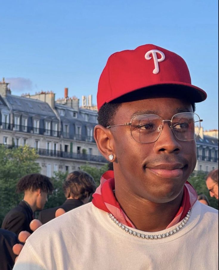 a man wearing a red hat and glasses standing in front of some buildings with other people