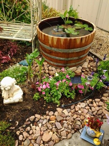 a wooden barrel filled with water surrounded by rocks and flowers in a garden area next to a bench