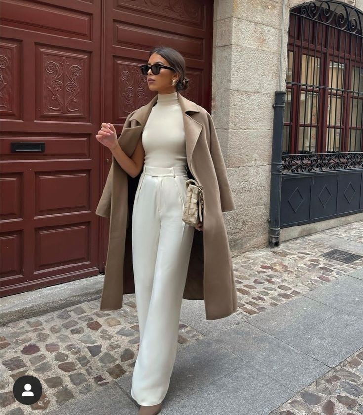 a woman standing in front of a red door wearing white pants and a tan coat