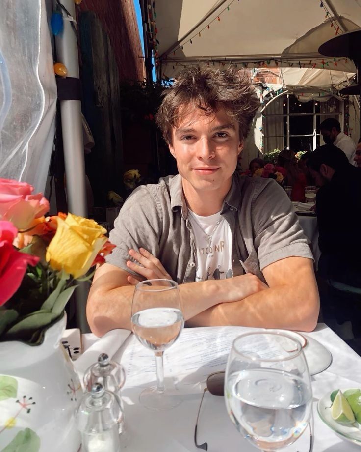 a young man sitting at a table with wine glasses and flowers in front of him