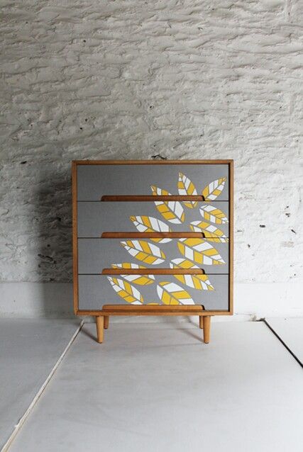 a dresser with yellow leaves painted on the front and sides, against a white brick wall